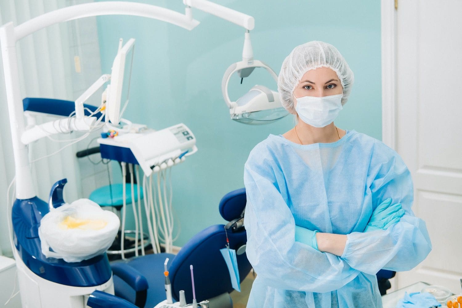 a female dentist wearing a medical mask and rubber gloves poses for the camera and folds her arms in her office.